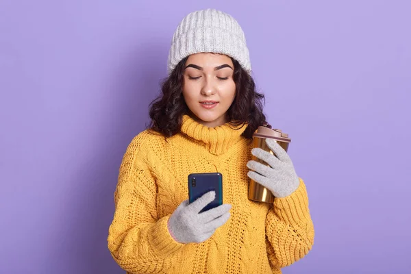 Képzeld el, ahogy egy csinos, elbűvölő, figyelmes nő szürke kalapot és kesztyűt visel, kezében okostelefonnal és termo bögrével, mindkét kezében forró itallal, figyelmesen nézi a képernyőt. Technológiai koncepció. — Stock Fotó