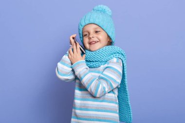 Horizontal indoor picture of adorable sweet good looking child being in good mood, holding smartphone near ear, looking aside, having conversation over phone. Children and technology concept.