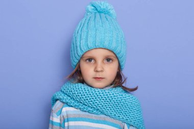 Indoor shot of little serious girl wearing cap, scarf and striped shirt standing isoalted over lilac background, looking dirtectly at camera with earnest facial expression, having negative emotions.