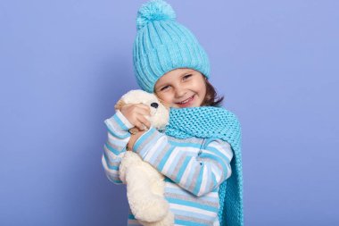 Horizontal photo of cute little girl dressed cap, scarf and stripes shirt, holding plush toys up to her face, looking directly at camera withcharming smile, having positive emotions. Chidhood concept.