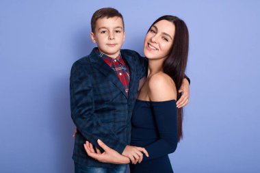 Portrait of delighted smiling young dark haired mother hugging her son, looking directly at camera, looking directly at camera, having pleasant facial expression. Family relationship concept.