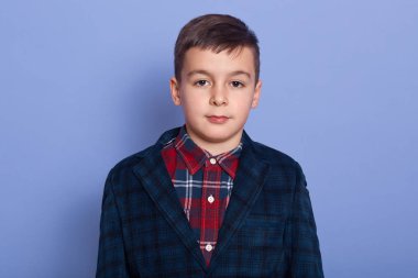 Close up portrait of little schoolboy looking directly at camera, standing straight isolated over blue background, wearing red plaid shirt and dark blue jacket, having peaceful facial expression.
