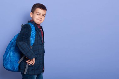 Indoor picture of funny cute thoughtful pupil going to school, being ready to study hard, having backpack with books aback, wearing dark blue jacket and plaid shirt. Copyspace for advertisement.