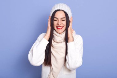 Image of emotional attractive young brunette standing isolated over blue background in studio, closing eyes, having toothy smile, wearing sweater and scarf, having red lips, putting hands on ears.