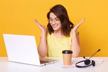 Studio shot of attarctive girl working with lap top while sitting at white desk, looking smiling at computer screen and spreading hands, isolated over yellow studio background. Technology concept.