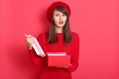 Valentine day concept. Sad woman opening empty box gift present. Elegant attractive model wearing red sweater and beret, looking directly at camera with upset facial expression. Studio shot.