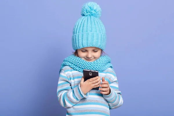 Retrato de niña adorable atenta con sombrero azul y bufanda, sudadera a rayas, de pie aislado sobre fondo lila, sosteniendo el teléfono inteligente en ambas manos, utilizando el dispositivo. Concepto técnico —  Fotos de Stock
