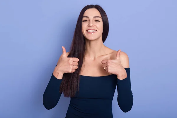 Portrait of attractive beautiful attractive young brunette standing isolated over lilac background, making gesture, showing sign of approval, raising thumbs up, wearing blue dress. Emotions concept. — Stockfoto