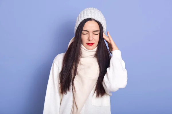 Horizontal picture of young attractive woman standing isolated over blue background in studio, putting hand on ear, having earache, having caught cold, closing eyes, wearing white warm clothes. — 图库照片