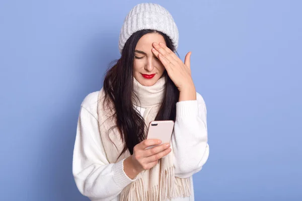 Horizontal shot of young girl with winter hat posing isolated over blue background, being unhappy and frustrated with something, holds smartphone.. Negative facial expression, touching her forehead. — Stock Photo, Image
