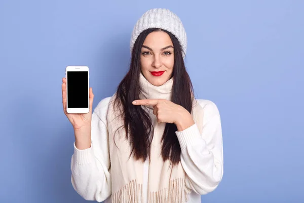 Студийный снимок молодой привлекательной женщины указывая на пустой экран телефона в руке и глядя прямо в камеру, Winsome леди с красными губами, одетые в теплый белый свитер и кепку, изолированные на синий . — стоковое фото
