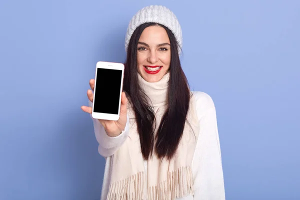 Крупный план портрет молодой женщины, показывающей смартфон с пустым экраном, Winsome леди одевается белый теплый свитер и зимняя кепка, позируя с красными губами изолированы на синем фоне студии. Копирование пространства . — стоковое фото