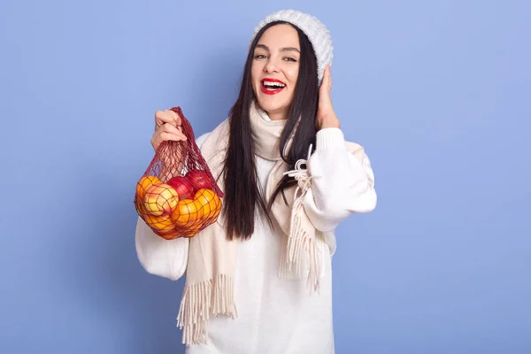 Horizontal shot of woman holding shopping bag full of apples and oranges, posing isolated over over blue background, lady dresses white clothes, looking smiling directly at camera with opened mouth. — Stockfoto
