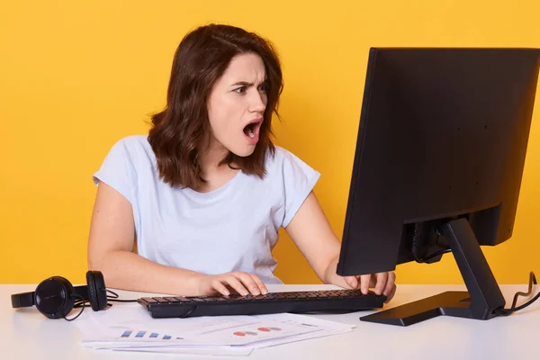 Horizontal shot of brunette woman using computer, reading shocking information on PC screen, posing with opened mouth isolated over yellow studio background, winsome lady wearing casual clothing. — Stock Photo, Image