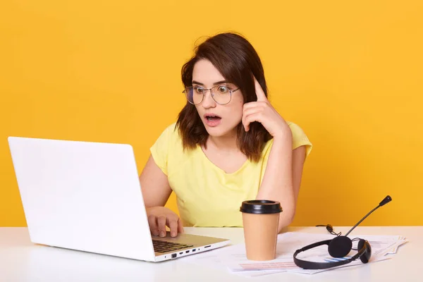 Οριζόντια λήψη της γυναίκας των επιχειρήσεων λειτουργεί σε απευθείας σύνδεση στο laptop της, ενώ κάθεται στο γραφείο, κοιτάζοντας την οθόνη με ευρέως ανοιγμένο στόμα, έχει εκπλαγεί και σοκαρισμένη έκφραση του προσώπου, απομονώνονται πάνω από κίτρινο τοίχο — Φωτογραφία Αρχείου