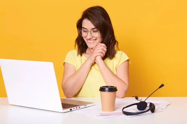 Foto wanita horisontal dengan laptop terisolasi di atas latar belakang kuning, wanita tersenyum yang menawan melihat monitor komputer, wanita yang minum kopi atau teh, menjaga tangan dekat wajah, mengungkapkan sukacita . — Stok Foto