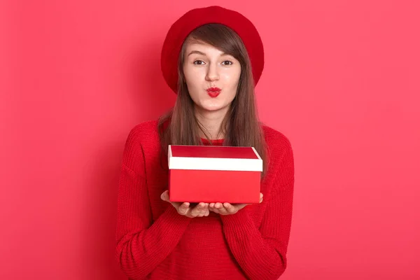 Крупный план портрет красивой женщины в красном свитере и берете, держа в руках красную подарочную коробку, делая жест поцелуя, хочет сделать подарок для своего парня, позируя изолированно над стеной студии . — стоковое фото