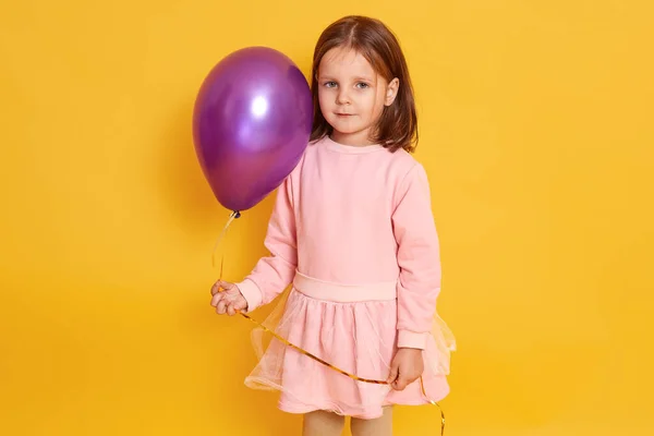 Quadro orizzontale interno di dolce positivo bella bambina in piedi isolato su sfondo giallo, tenendo palloncino d'aria in una mano, guardando direttamente la fotocamera, indossando abito rosa . — Foto Stock