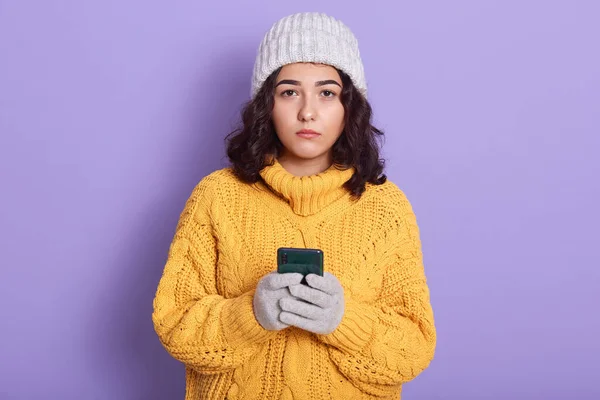 Fotografía interior horizontal de morena decepcionada mirando directamente a la cámara, de pie aislada sobre fondo lila, sosteniendo el teléfono inteligente en ambas manos, usando ropa de abrigo. Concepto técnico . — Foto de Stock