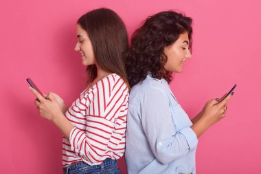 Pembe arka planda yalnız poz veren iki güzel kadının yakın plan portresi. Sırt sırta verip duran güzel esmer kadınlar gündelik kıyafetler giyiyorlar. Holding telefonları..