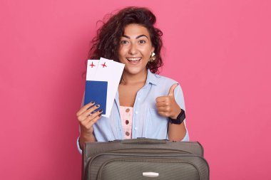 Kapalı alanda çekilen genç, çekici bir turist kadının günlük giysiler giydiği, bavul, bilet ve pasaport ile poz verdiği, pembe duvarın üzerinde izole bir şekilde baş parmağını gösterdiği bir fotoğraf. Yurt dışına seyahat eden yolcu.