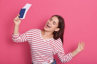 Pasaport ve uçuş kartı taşıyan neşeli beyaz kadın turistin pembe stüdyo arka planında izole edilmiş pozu mutlu görünüyor, kız mutlu şarkı söylüyor..