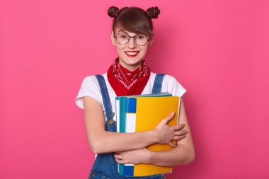 Renkli kâğıt klasörlü, bandana, gömlek ve tulum giyen, kameraya bakıp gülümseyen, komik saçlı, pembe arkaplanda izole bir şekilde poz veren mutlu bir öğrencinin portresi..