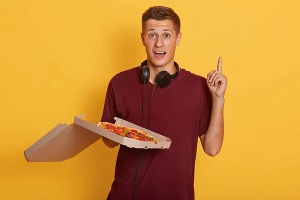 Tiro horizontal del repartidor de pizza con ropa casual sosteniendo cajas de pizza sobre un fondo amarillo aislado con el dedo índice hacia arriba, teniendo buena idea, chico guapo con expresión fasial asombrosa — Foto de Stock