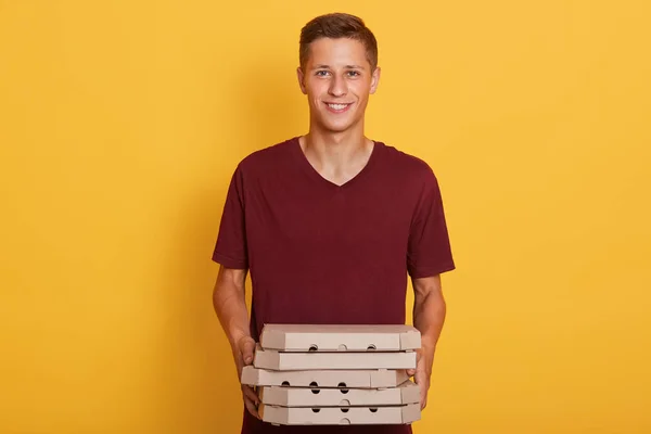 Photo hirizontale d'un bel homme portant un t-shirt décontracté marron, tenant des boîtes à pizza dans les mains, regardant directement la caméra, posant isolé sur un fond de studio jaune. Concept de livraison et de service . — Photo