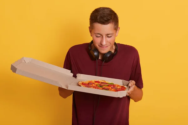 Primer plano retrato de chico guapo con camiseta borgoña oliendo pizza fresca de caja de cartón y obtiene placer, de pie aislado sobre el fondo amarillo, joven que está listo para comer comida chatarra . — Foto de Stock