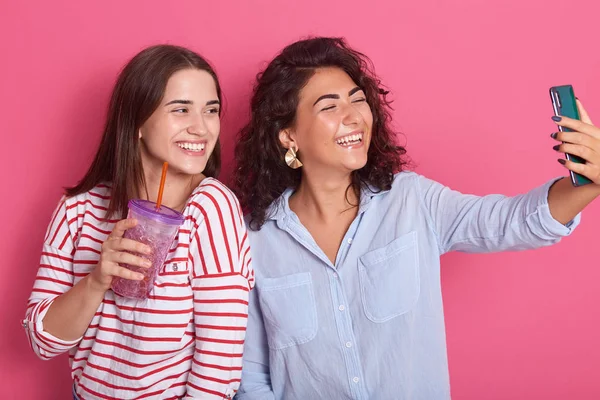 Disparo horizontal de dos amigas chicas tomando selfie con teléfono inteligente, posando aislado sobre fondo rosa, dama lleva camisa casual con la botella de aguada manos, mirada feliz, expresando felicidad — Foto de Stock