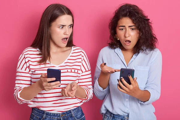Picture of two surprised cute young women pose together isolated over pink background, brunette females with widely opened mouths, look astonished, wearing casual clothing, checking networks. — Stock Photo, Image