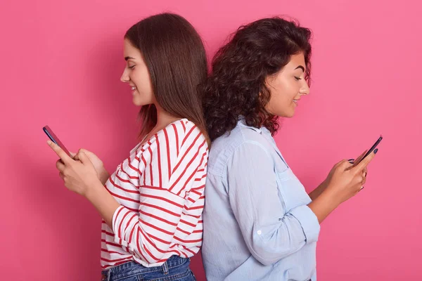 Close up portrait of two cute young women pose together isolated over pink background while standing back to back to each other, beautiful brunette females wearing casual clothing, holdings phones. — Stock Photo, Image