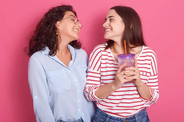 Indoor shot de dos chicas adultas atractivas de pie en el estudio, vistiendo ropa casual, mirándose sonrientes, posando aisladas sobre un fondo rosa, las mujeres expresando felicidad y alegría . — Foto de Stock