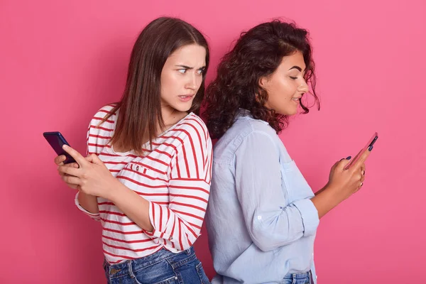 Beltéri felvétel nőkről, akik mobiltelefonokat néznek, és rossz híreket vagy fotókat látnak, miközben egymásnak háttal állnak. Emberi érzelem, reakció, kifejezés, pózol elszigetelt rózsaszín fal. — Stock Fotó