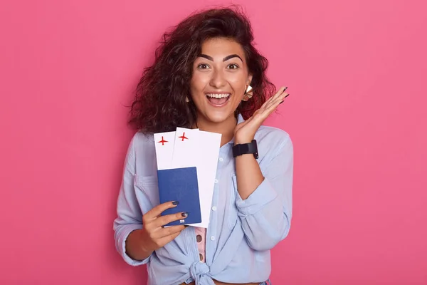 Horizontal shot of beautiful modern smiling woman with passport and air tickets in her hand, lady wearing stylish clothing, model posing isolated over pink background, looks surprised and happy.
