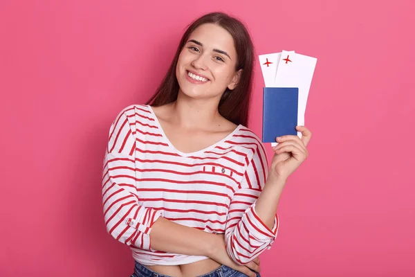 Half length portrait of female tourist holding airline tickets and passport, modeling posing isolated over pink studio background, female wearing stripped shrt, looking at camera with charming smile.