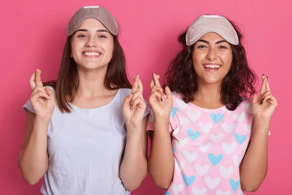 Kép két fiatal vidám nők ruhák elegáns pizsama és bekötött szemek, állni mosolygós és tartja ujjak keresztbe elszigetelt rózsaszín stúdió háttér, nézi a kamera, kifejező boldogság. — Stock Fotó