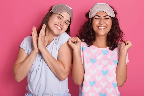 Indoor shot of lovely young European women with pleasant smile, wearing blindfolds and pajamas, standing isolated over pink studio wall, expresses good emotions, females having good time at home. — Stock Photo, Image