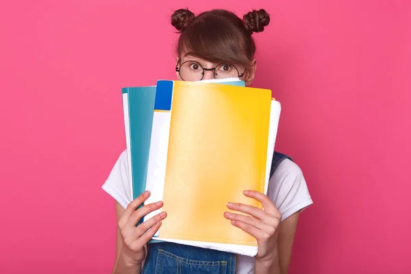 Studio shot of emotive european woman hides behind blue and yellow paper folders, wearing round glasses, white t shirt, denim overalls, standing against pink background, girlhaving funny hair buns.