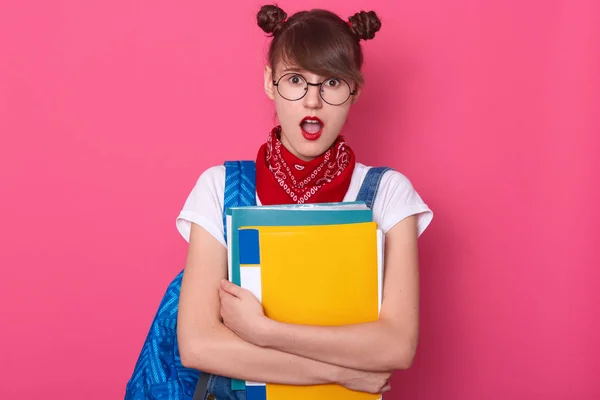 Primer plano retrato de estudiante con dos racimos divertidos, usando camiseta blanca, overoles de mezclilla, pañuelo rojo y gafas redondeadas, sosteniendo la carpeta de papel, ha conmocionado la expresión facial, posando con la boca abierta — Foto de Stock