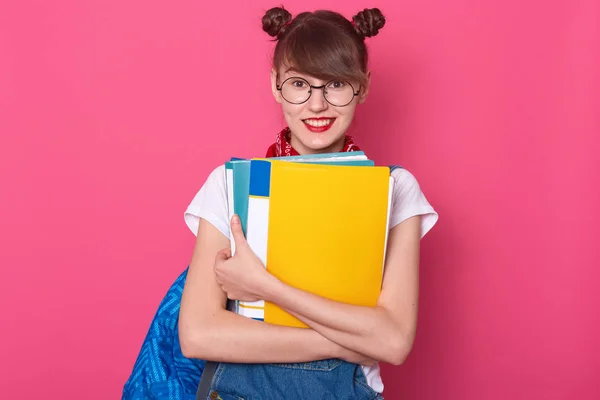 年轻的黑发白人学生女孩戴着眼镜，白色T恤，拿着纸夹和蓝色背包，站在粉色背景上的迷人女性形象。教育概念. — 图库照片