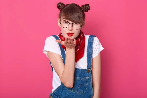 Close-up portret van aantrekkelijke donker haar meisje blazen luchtkus rechtstreeks naar de camera, strekt de hand, heeft een aantrekkelijke look, gekleed wit t-shirt, denim overalls, poseren geïsoleerd over roos achtergrond. — Stockfoto