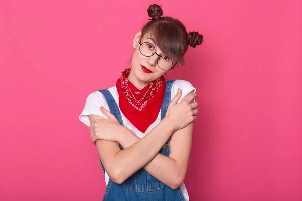 Close-up portret van aantrekkelijke vrouw dragen van denim overalls, wit casual t-shirt, rode bandana over de hals, poseren geïsoleerd over roze achtergrond, vrouw kijkt direct naar de camera, knuffelen zichzelf. — Stockfoto