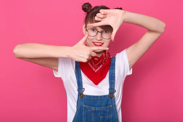 Horizontal tiro de sorrir adolescente framing rosto com os dedos, fingindo capturar foto, posando isolado sobre o fundo do estúdio rosa, estudante menina vestidos macacão, tendo pães de cabelo engraçado . — Fotografia de Stock