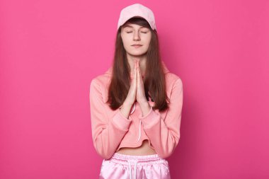 Huzurlu genç bir kadının portresi avuç içlerini birleştiriyor, gözlerini kapatıyor, uzun kahverengi saçlara sahip, pembe kapüşonlu, pantolonlu ve şapkalı, yalnız dua eden, sessizliğini koruyan. Barış kavramı.