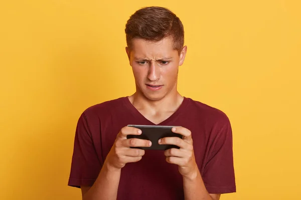 Retrato de guapo curioso entretenido joven de pie aislado sobre el fondo amarillo, sosteniendo el teléfono inteligente en ambas manos, estar ocupado, jugar juegos en línea. Concepto de personas y tecnologías . — Foto de Stock