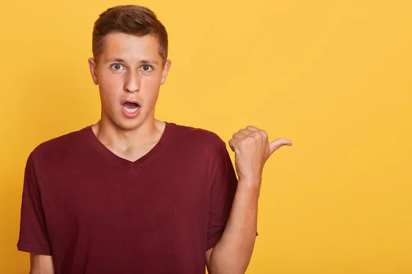 Retrato de un joven emocionalmente guapo mirando, mostrando dirección, haciendo gesto, con el pulgar hacia arriba, usando una camiseta de color rojo oscuro, abriendo ampliamente la boca y los ojos. Copyspace para publicidad . — Foto de Stock