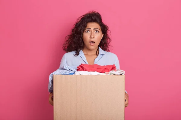 Portrait of shocked emotional young female standing isolated over pink background in studio, opening mouth widely, having surprised facial expression, having carton box with donated clothes in hands. — Stock Photo, Image