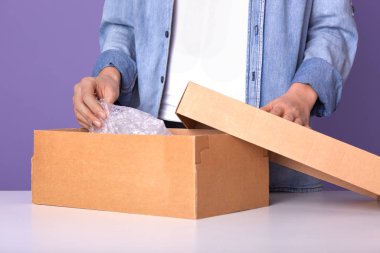 Kot ceket ve beyaz gömlek giyen adamın suratsız portresi, karton kutu paketi açan adam, beyaz masada hediye taşıyan erkek eflatun rengi arka planda izole edilmiş poz veriyor. Teslimat konsepti.